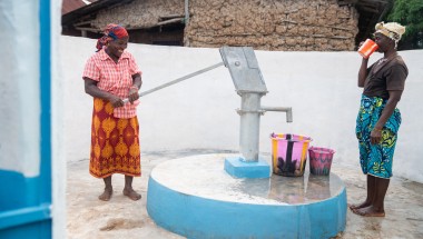Two individuals stand at a freshly painted water pump within a walled outdoor area. One person operates the hand pump, while the other drinks from a cup. Several colourful buckets are placed on the ground near the pump, and a building made of natural materials is visible in the background.