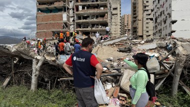Aid worker and resident stand beside rubble as rescue teams search debris in front of a heavily damaged apartment building.