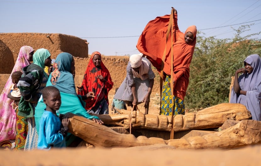 Women collect water from a wooden village well. Her orange hijab billows in the wind as she pulls water up from the well. Behind are orange building and a clear, sunny sky.