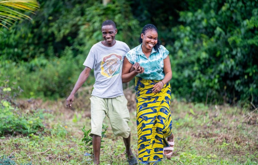  Two people walk, smiling together through a grassy, open area bordered by dense green vegetation. One person wears a grey T‑shirt and light shorts, while the other wears a patterned yellow and blue skirt and a teal top. They hold onto each other’s arms as they move forward across the uneven ground.