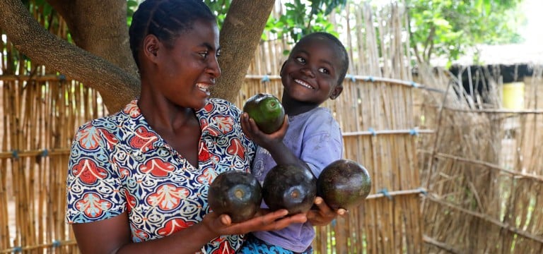 A smiling woman holds several avocados in her hands while carrying a young child, who happily holds up another avocado. They stand under a tree beside a wooden fence.