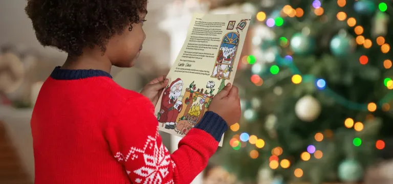 A young child in a red Christmas jumper reads a colourful letter from Santa while sitting by a decorated Christmas tree with glowing lights and ornaments.