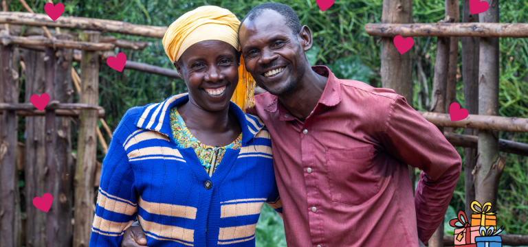 A smiling couple stands close together outdoors, with the man’s arm around the woman’s shoulders. They are surrounded by greenery and a wooden fence, with pink heart graphics and a small gift illustration added around them, creating a warm, celebratory feel.