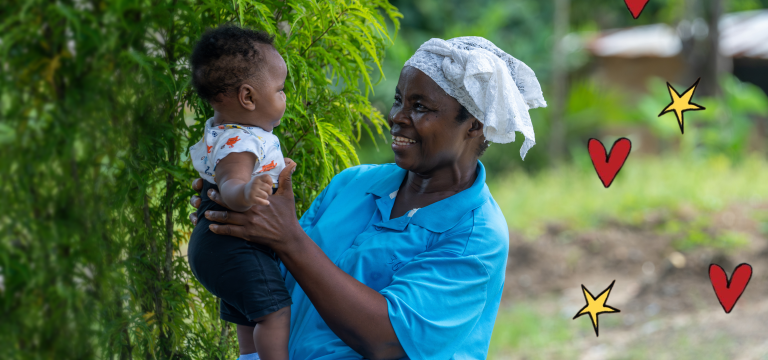 An older woman wearing a blue top and white headscarf sits outdoors holding a baby, smiling as they look at each other. Green foliage surrounds them, with a rural setting and a small building softly blurred in the background.