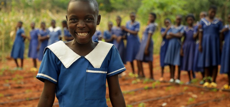 A smiling schoolgirl wearing a blue school uniform stands in the foreground of a school garden. Behind her, a group of students in matching blue uniforms stand together, slightly out of focus. Rows of young plants grow in the red soil around them, with green vegetation in the background. The image conveys education, community, and young people participating in school-based gardening activities.
