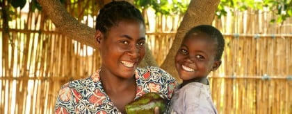 A smiling woman holds several large avocados while standing beside a young boy in her arms, who is also holding an avocado. Both are beaming with joy, standing under the shade of a tree with a bamboo fence in the background.