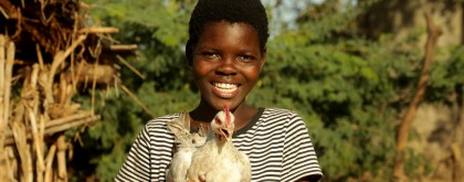 A smiling young person stands outdoors holding a white chicken in both hands. Trees and simple wooden structures are visible in the background, suggesting a rural setting.