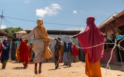 A group of children and young people stand on sandy ground in an outdoor area, skipping with ropes and playing together. Two figures in the foreground swing skipping ropes, while others gather and watch near corrugated metal buildings and structures with tin roofs under a bright blue sky.