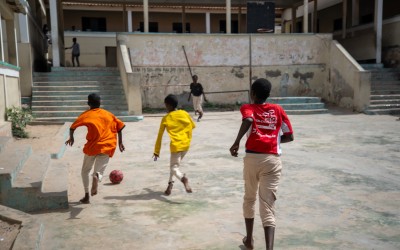Three children run across a concrete courtyard, chasing a red-and-black football toward another child near a wall. The scene takes place in a worn school-like courtyard with faded walls, steps on both sides, and a multi-level building surrounding the area.