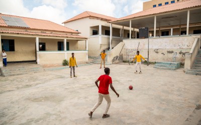 Four young people stand and move around a concrete courtyard, playing with a red-and-black football. One person in a red shirt approaches the ball in the foreground, while three others in yellow shirts stand spaced out facing them. The courtyard is bordered by weathered school buildings with peeling paint, steps on both sides, and a covered walkway supported by columns, with solar panels visible on one roof.