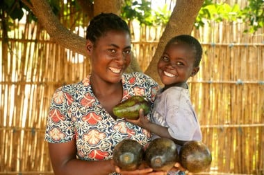 A smiling woman and child stand outdoors holding several large avocados, with a bamboo fence in the background.