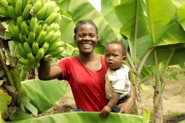 Smiling woman in a red shirt holds a baby in one arm and a large bunch of green bananas in the other, standing among banana trees.