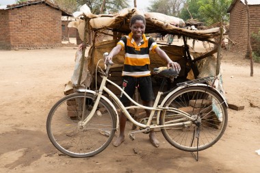 Smiling boy in a striped shirt stands barefoot outdoors, proudly holding a bicycle in front of a small makeshift shelter.