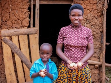 Smiling woman and young boy stand outside a mud-brick home, proudly holding fresh eggs in their hands.