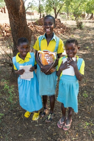 hree smiling schoolchildren in blue and yellow uniforms stand outdoors, holding notebooks and a school bag.