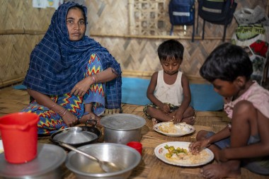 A woman in a blue headscarf sits on the floor of a bamboo shelter with two children eating rice and vegetables from plates.