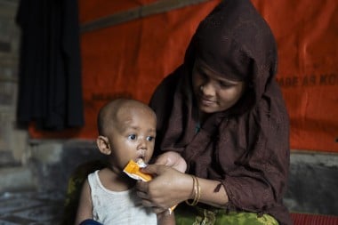 A woman in a brown shawl carefully feeds a young child therapeutic food paste from a packet.
