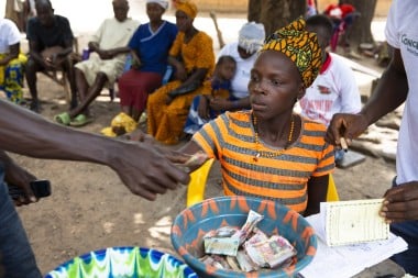 A woman wearing an orange and grey striped dress and patterned headscarf collects money into a colorful plastic bowl during a community meeting. Several people sit in the background under the shade of trees, some observing while others hold notebooks or cash.