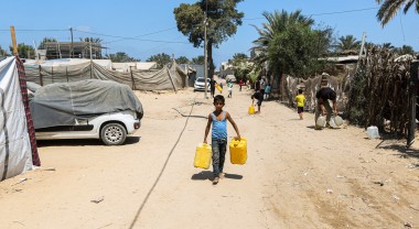A young boy walks barefoot along a sandy road carrying two large yellow water containers, with makeshift shelters and covered cars lining the path. Other people and children in the background also carry water containers under the bright midday sun.