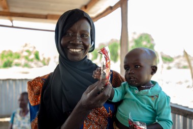 A smiling woman wearing a black headscarf holds a baby in her arms, both holding packets of nutritional food. They stand under a shaded structure with sunlight filtering through, and other people are visible in the background.