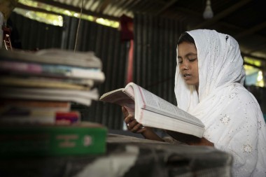 A young woman wearing a white headscarf sits indoors reading a large open book with focus and concentration. A stack of books lies on the table beside her, and soft daylight filters through the corrugated metal walls of the room.
