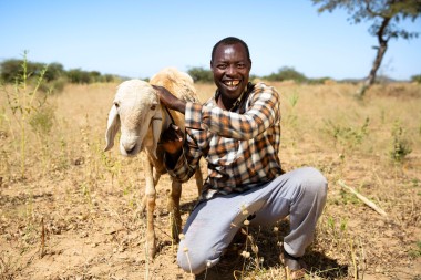 A man kneels on dry, grassy land, smiling broadly while gently holding a white goat beside him. He wears a plaid shirt and grey trousers, and the sunny background shows sparse vegetation and a clear blue sky.