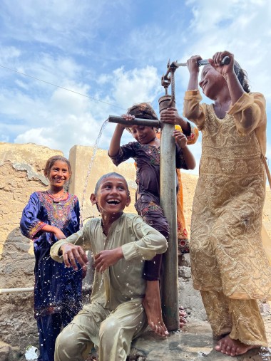 Four smiling children play joyfully around a water pump, with one operating the handle as clean water splashes out. The children, dressed in colourful traditional clothes, laugh under the bright daylight against a simple wall and blue sky background.