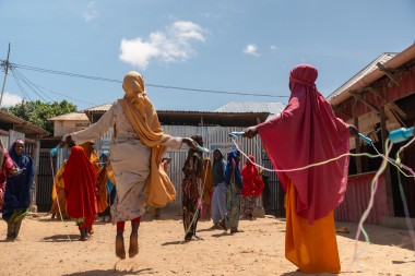 A group of children and young people stand on sandy ground in an outdoor area, skipping with ropes and playing together. Two figures in the foreground swing skipping ropes, while others gather and watch near corrugated metal buildings and structures with tin roofs under a bright blue sky.