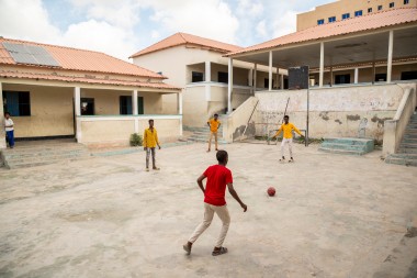 Four young people stand and move around a concrete courtyard, playing with a red-and-black football. One person in a red shirt approaches the ball in the foreground, while three others in yellow shirts stand spaced out facing them. The courtyard is bordered by weathered school buildings with peeling paint, steps on both sides, and a covered walkway supported by columns, with solar panels visible on one roof.