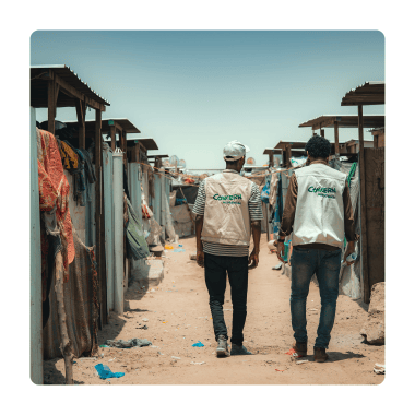 Two men wearing Concern branded gilets walk away together on a dusty path surrounded by temporary shelters.