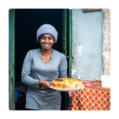 A smiling woman wearing a grey knit hat stands in the doorway of her home, holding a freshly baked round cake on a serving plate. A small table with kitchen containers sits beside her, and the simple home setting creates a warm, welcoming atmosphere.