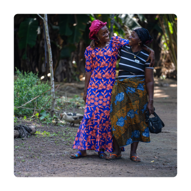 Two smiling women walk arm in arm along a village path, looking warmly at one another. Surrounded by lush tropical greenery, they wear colourful patterned clothing, conveying friendship, joy and mutual support.