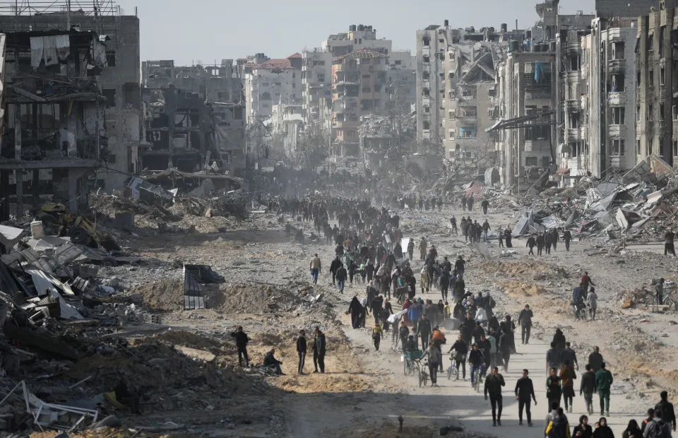 Crowds of people walk through a wide street filled with rubble and debris, flanked by heavily damaged and destroyed buildings in a war-torn city.