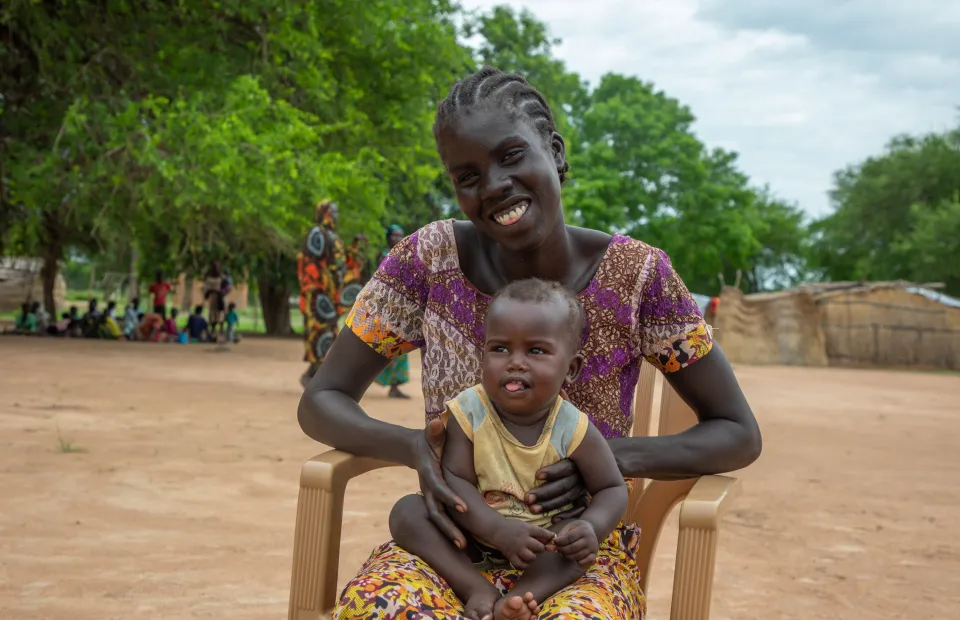 A mother sits on a charity with her happy baby on her lap. Behind her are people in colourful clothing and large green trees. 