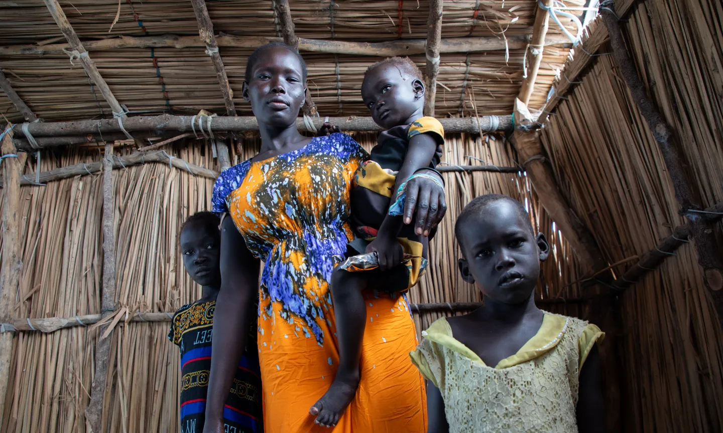 A woman stands inside a shelter made of sticks, holding a small child on her arm. Two other children stand close beside her, looking directly at the camera. The woman wears a brightly patterned orange and blue dress, and the children are dressed in colorful clothes.