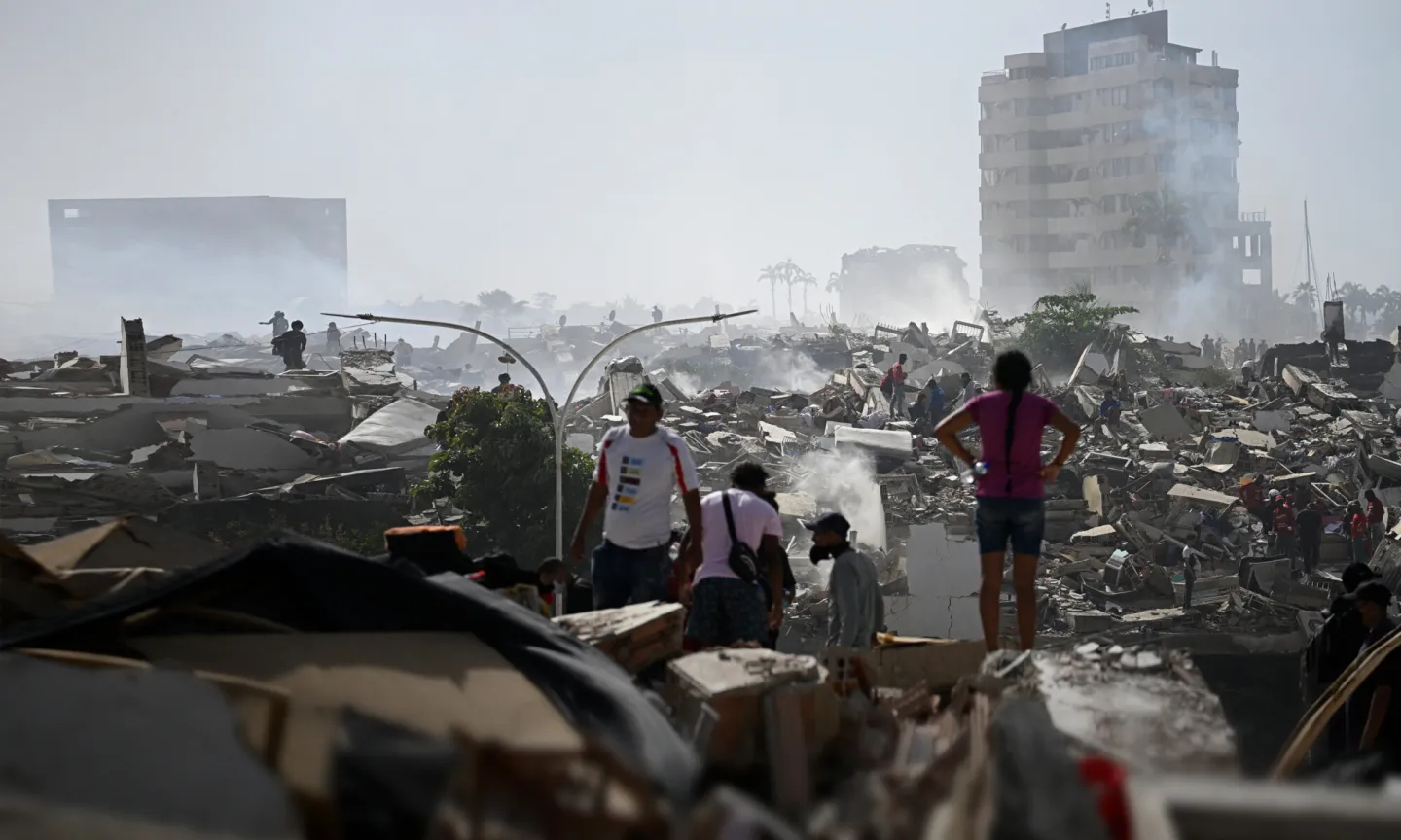 People walk through the aftermath of widespread destruction in a heavily damaged urban area, where collapsed buildings, debris, and smoke stretch across the landscape. A few damaged high-rise buildings remain standing in the background as residents and responders move carefully through the ruins.