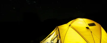 A glowing yellow tent illuminated from within stands in a dark campsite under a star-filled night sky. Hiking boots and trekking poles rest outside the entrance, suggesting a day of adventure.