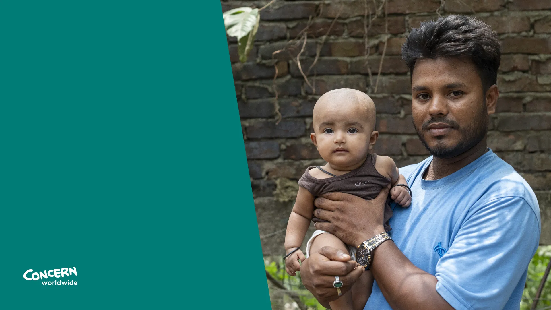 A man in a blue shirt holds a baby in his arms while standing outdoors in front of a brick wall. The baby looks directly at the camera, and both appear calm and composed.