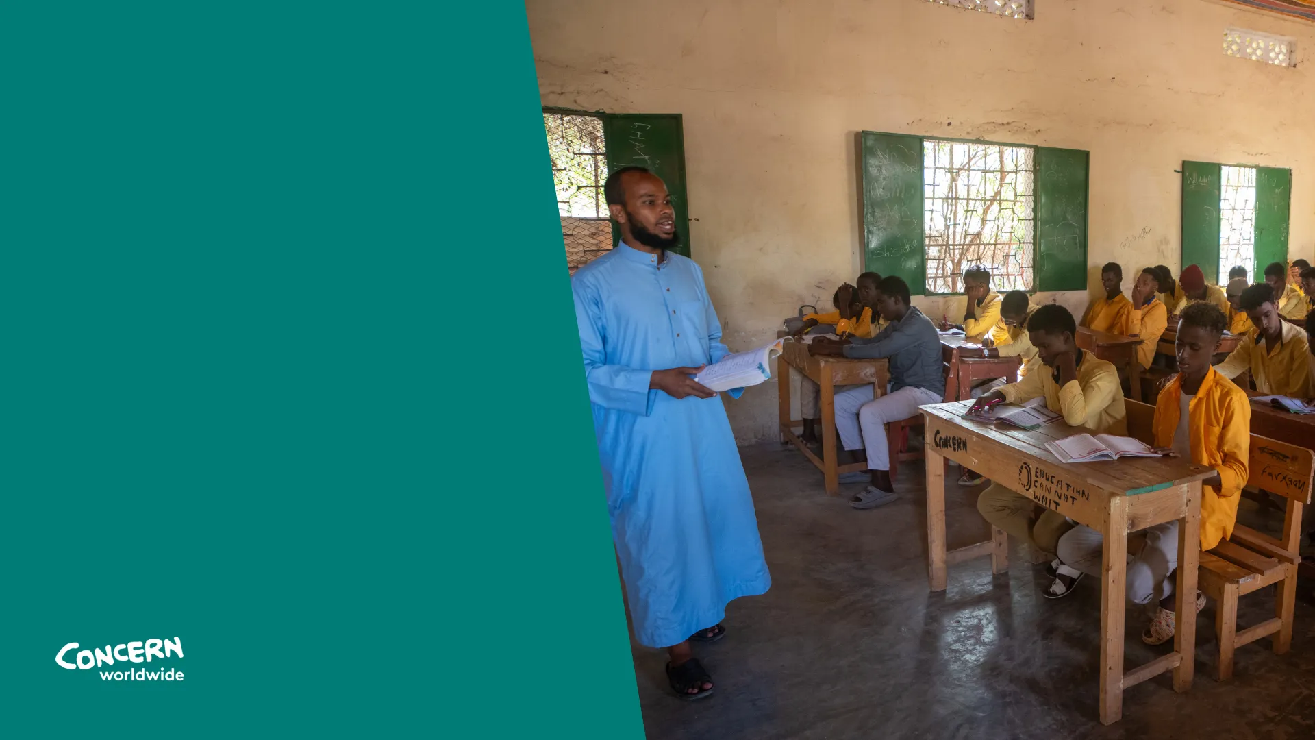A teacher in a light blue robe stands at the front of a classroom, holding a book and addressing a group of students. The students, dressed mostly in yellow uniforms, sit at wooden desks with open notebooks, attentively following the lesson in a brightly lit classroom with green window shutters.