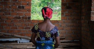 A woman in a colourful dress and headscarf looks out of a window in a brick building.