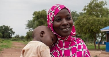 A person wearing a pink and white patterned headscarf holds a child outdoors. In the background, there are trees and a building with a blue roof.