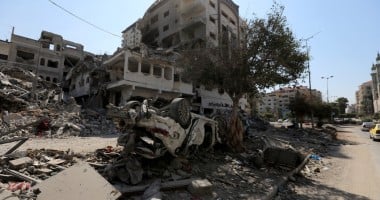 Scene of destruction with collapsed buildings and scattered debris. An overturned car is partially buried under rubble. Damaged structures and a lone tree are visible in the background under a clear daytime sky.