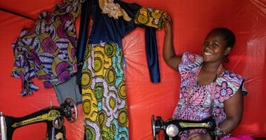 Woman smiling while seated at a sewing machine, holding up a colorful dress with yellow, blue, and green patterns and lace detailing. A second garment with purple and pink designs hangs nearby against a red fabric backdrop