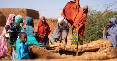 Women collect water from a wooden village well. Her orange hijab billows in the wind as she pulls water up from the well. Behind are orange building and a clear, sunny sky.