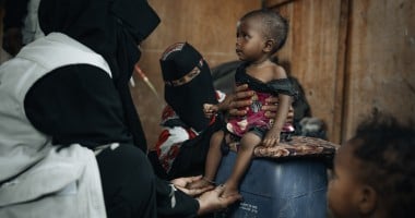  healthcare worker dressed in black clothing with a white vest is examining a small child seated on a blue plastic container. Another person wearing patterned clothing and a black face covering supports the child. The child is wearing a red and black outfit and appears thin, with visible ribs and limbs. The setting looks modest, with a brown wall in the background and another child partially visible in the foreground.