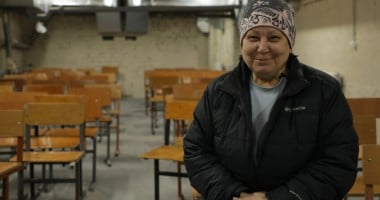 A person wearing a winter jacket and patterned hat sits in a room filled with rows of wooden school chairs. The setting appears to be a basement shelter with exposed brick walls and overhead ventilation ducts.