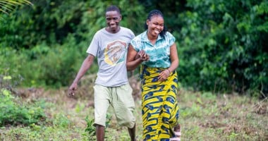  Two people walk, smiling together through a grassy, open area bordered by dense green vegetation. One person wears a grey T‑shirt and light shorts, while the other wears a patterned yellow and blue skirt and a teal top. They hold onto each other’s arms as they move forward across the uneven ground.