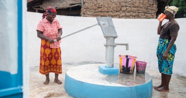 Two individuals stand at a freshly painted water pump within a walled outdoor area. One person operates the hand pump, while the other drinks from a cup. Several colourful buckets are placed on the ground near the pump, and a building made of natural materials is visible in the background.