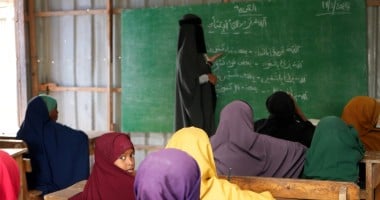 Teacher writing on chalkboard while children sit at desks in a classroom.