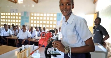 A young woman in a classroom standing beside a microscope.